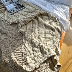 May include: A cream-colored throw blanket with brown stripes is folded on a white bed. The blanket has a textured, waffle-like weave.