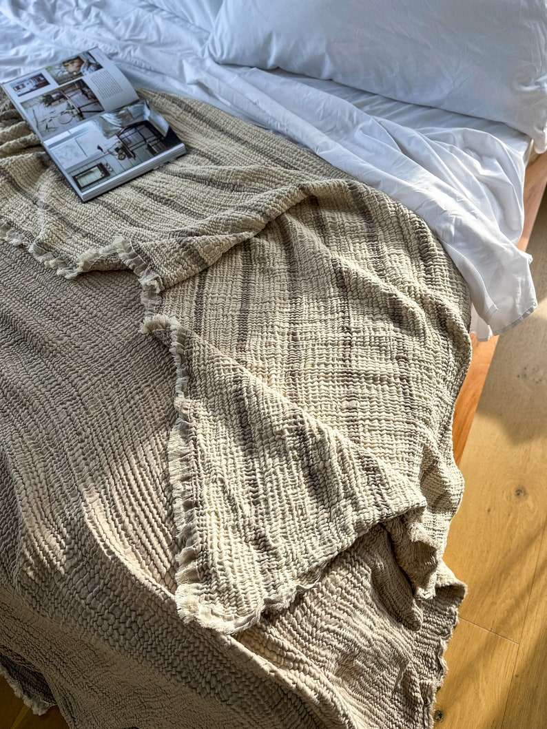 May include: A cream-colored throw blanket with brown stripes is folded on a white bed. The blanket has a textured, waffle-like weave.