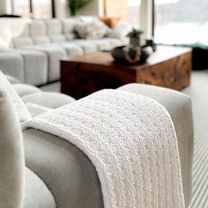 May include: A white, textured throw blanket with a fringed edge draped over a gray armchair. The blanket's intricate pattern and soft texture are visible. The background shows a modern living room with a large sectional sofa and a wooden coffee table.