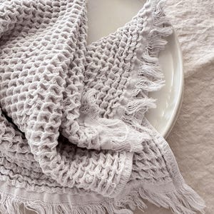 May include: A light gray waffle weave towel with a fringed edge. The towel is folded and draped over a white plate.