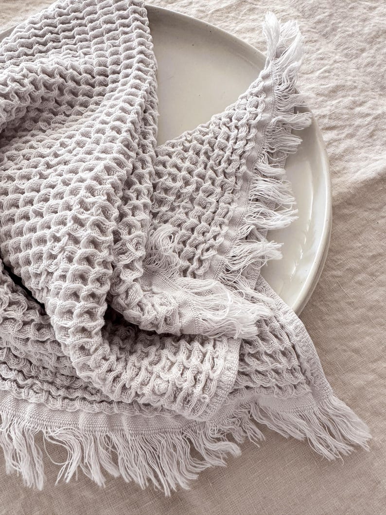 May include: A light gray waffle weave towel with a fringed edge. The towel is folded and draped over a white plate.