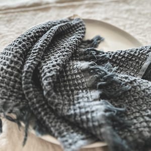 May include: A dark gray waffle weave kitchen towel with frayed edges. The towel is folded and placed on a white plate.