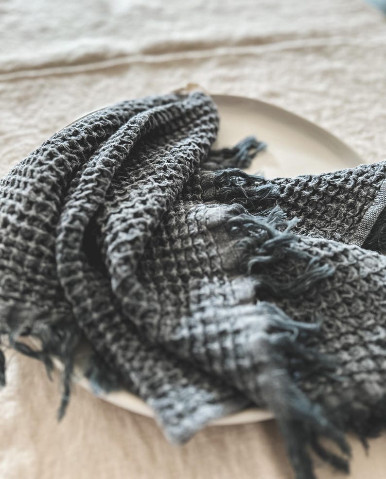 May include: A dark gray waffle weave kitchen towel with frayed edges. The towel is folded and placed on a white plate.