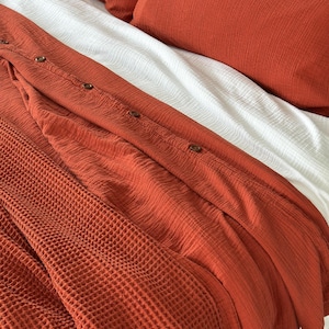 May include: Close-up of a bed with rust-colored bedding, including a button-down shirt, a waffle-knit blanket, and a pillow. White sheets provide contrast. The textures of the fabrics are visible, creating a cozy and inviting aesthetic.