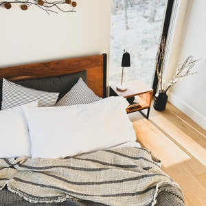 May include: A bed with a gray and white striped throw blanket, white pillows, and a wooden headboard. The bed is in a bedroom with a window overlooking a snowy forest.