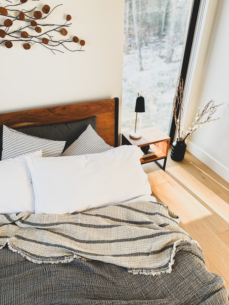 May include: A bed with a gray and white striped throw blanket, white pillows, and a wooden headboard. The bed is in a bedroom with a window overlooking a snowy forest.