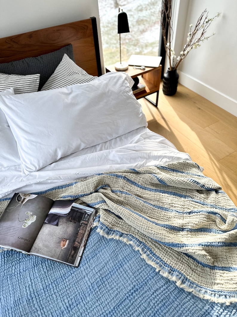 May include: A blue and white striped throw blanket with a fringed edge is draped over a white bed with a white pillow and a magazine on top. The bed is in a bedroom with a wooden headboard and a nightstand with a lamp.