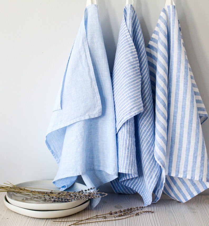 May include: Three blue kitchen towels hanging on hooks. Two towels have a white stripe pattern, and one towel is a solid light blue color.