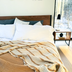 May include: A close-up of a bed with a white and brown striped throw blanket on top of a mustard yellow quilted blanket. The bed has a wooden headboard and two white pillows.