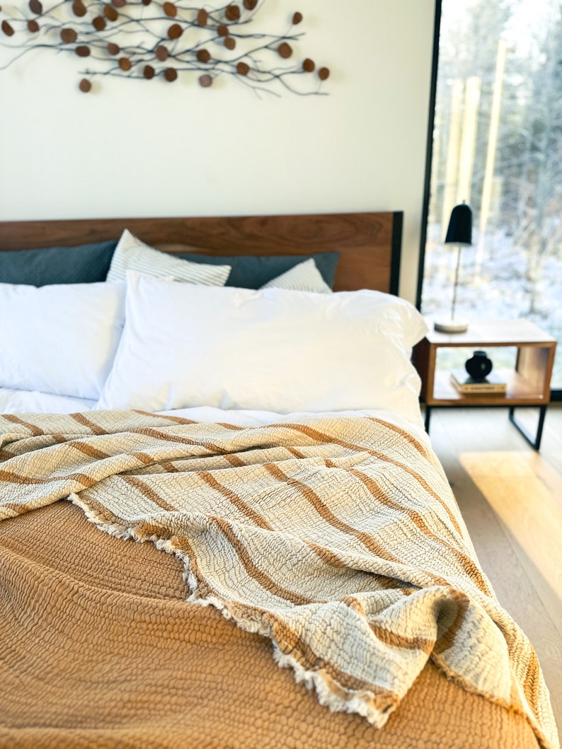 May include: A close-up of a bed with a white and brown striped throw blanket on top of a mustard yellow quilted blanket. The bed has a wooden headboard and two white pillows.