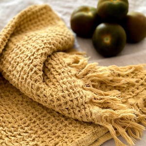 May include: A yellow waffle weave towel with fringed edges. The towel is folded and lying on a light beige surface.