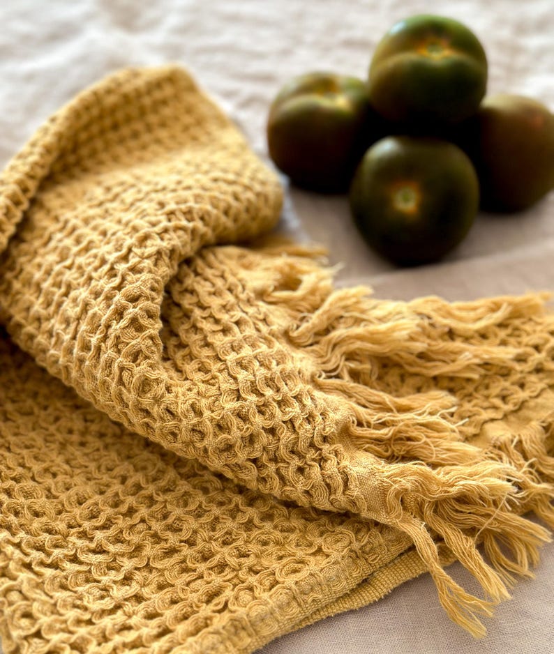 May include: A yellow waffle weave towel with fringed edges. The towel is folded and lying on a light beige surface.