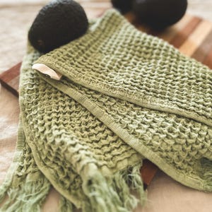 May include: A green waffle weave kitchen towel with fringe edges. The towel is folded and lying on a wooden cutting board.