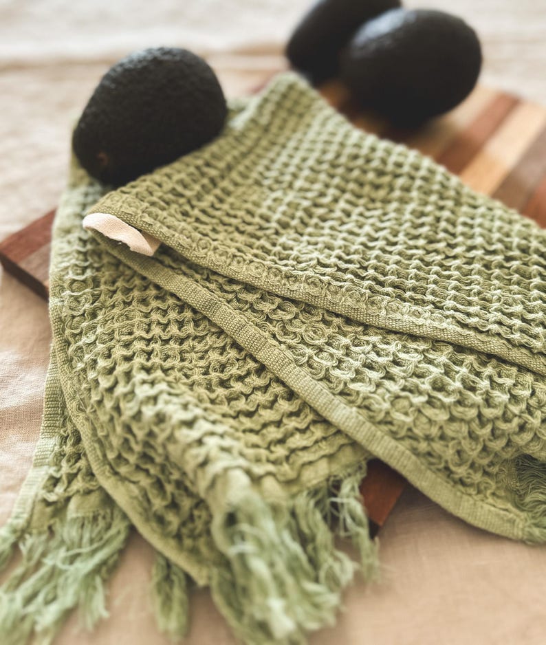 May include: A green waffle weave kitchen towel with fringe edges. The towel is folded and lying on a wooden cutting board.