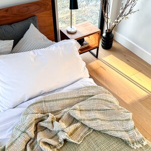 May include: A white bed with a green and white striped throw blanket. The bed has a wooden headboard and a white pillow. A black lamp is on a wooden nightstand next to the bed.