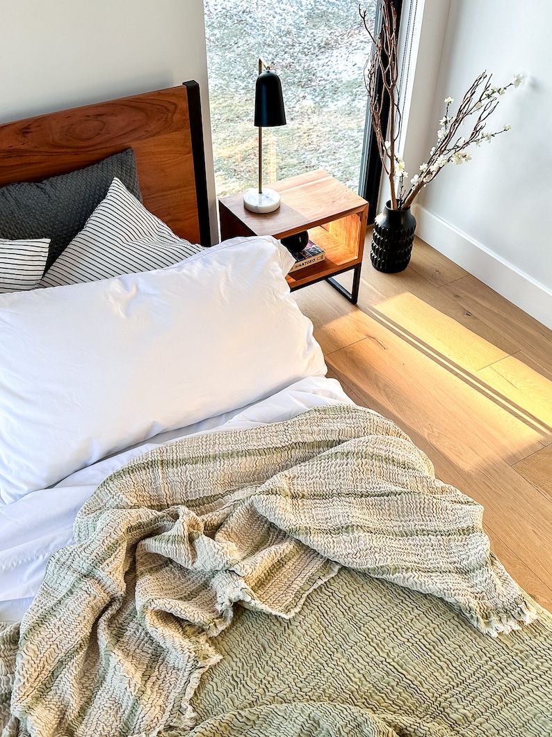 May include: A white bed with a green and white striped throw blanket. The bed has a wooden headboard and a white pillow. A black lamp is on a wooden nightstand next to the bed.