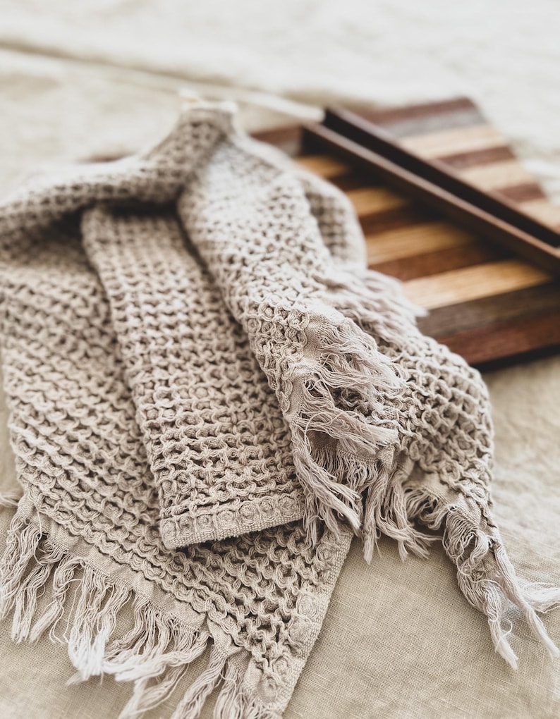 May include: A beige waffle weave towel with fringe edges. The towel is folded and draped over a wooden surface.