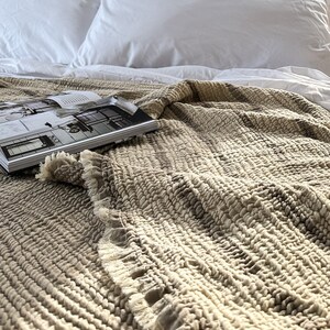 May include: A close-up of a beige textured throw blanket with a fringed edge. The blanket is folded and draped over a white bed with white pillows.