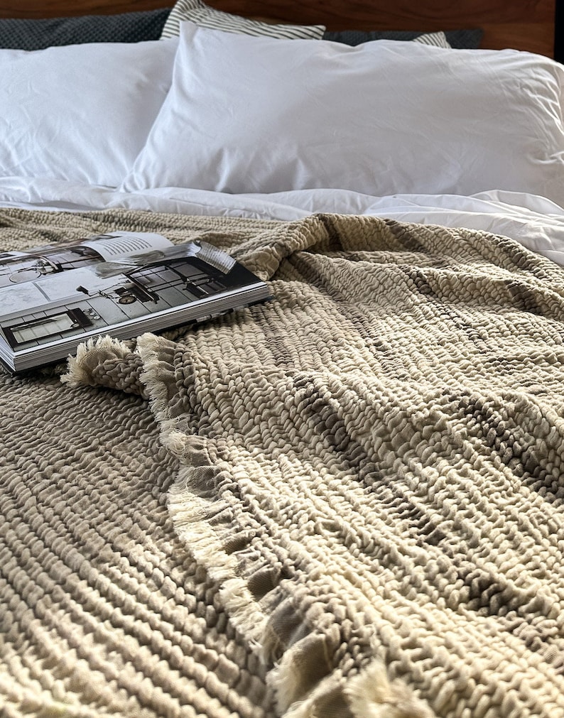 May include: A close-up of a beige textured throw blanket with a fringed edge. The blanket is folded and draped over a white bed with white pillows.