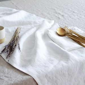 May include: A white linen table runner with a white ceramic cup and dried lavender sprigs on a beige linen tablecloth. Gold flatware is arranged on a white linen napkin.