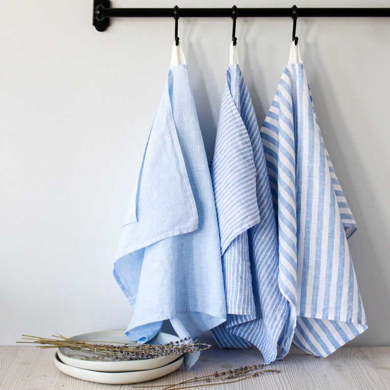 May include: Three blue and white striped kitchen towels hanging on a black metal rod. The towels are made of linen and have a subtle texture.