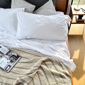 May include: A white bed with a wooden headboard, white bedding, and a striped throw blanket. There are two pillows with black and white stripes and one gray pillow on the bed. A book is lying on the blanket.