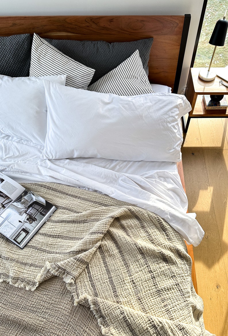 May include: A white bed with a wooden headboard, white bedding, and a striped throw blanket. There are two pillows with black and white stripes and one gray pillow on the bed. A book is lying on the blanket.