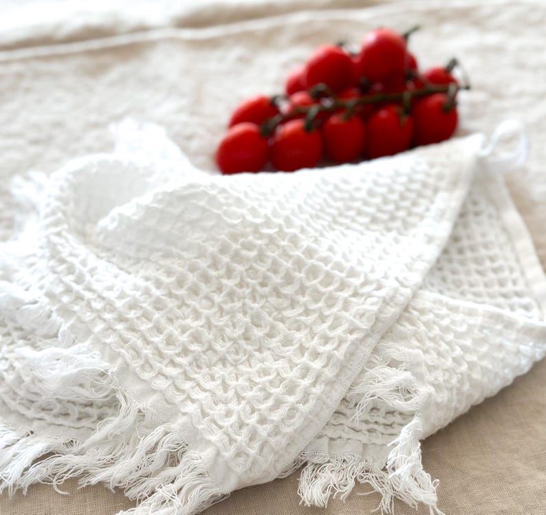 May include: White waffle weave kitchen towel with fringe edges. The towel is folded in half and has a textured waffle pattern.