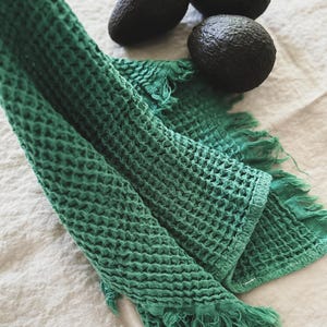 May include: A green waffle weave kitchen towel with fringed edges. The towel is folded and draped over a white linen surface. Three avocados are in the background.