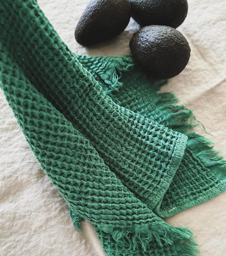 May include: A green waffle weave kitchen towel with fringed edges. The towel is folded and draped over a white linen surface. Three avocados are in the background.