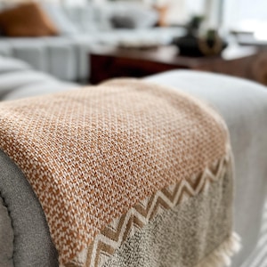 May include: A close-up of a brown and cream throw blanket with a geometric pattern. The blanket is draped over a gray couch.