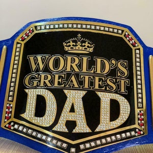 May include: A blue and gold championship belt with a black center plate that reads "WORLD'S GREATEST DAD" in gold lettering with rhinestones. The belt has a gold crown on top of the plate and red rhinestones on the sides.