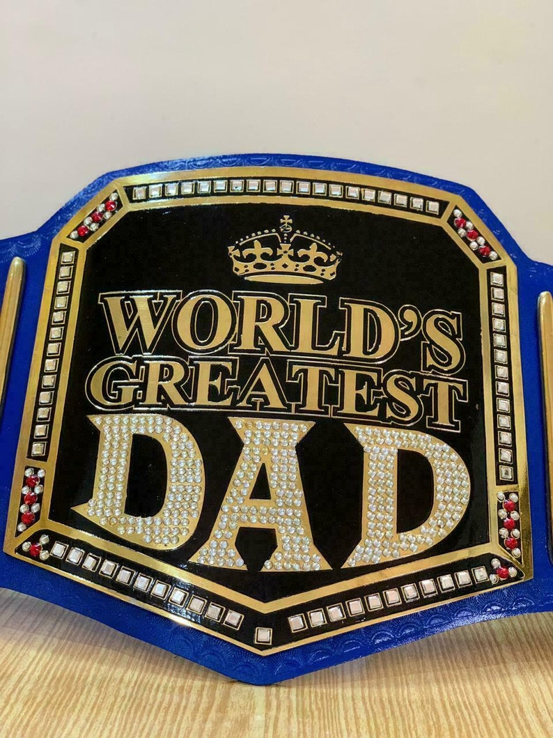 May include: A blue and gold championship belt with a black center plate that reads "WORLD'S GREATEST DAD" in gold lettering with rhinestones. The belt has a gold crown on top of the plate and red rhinestones on the sides.