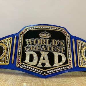 May include: A blue and gold championship belt with rhinestones. The center plate reads "World's Greatest Dad" with a crown above the text. The side plates read "Best Dad Ever".
