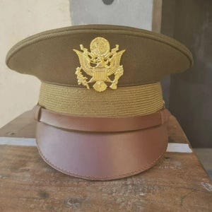 Pode incluir: Boné de serviço militar verde-oliva com um emblema dourado e uma viseira de couro castanho. O boné apresenta uma faixa dourada ao redor da coroa. Um chapéu militar clássico.