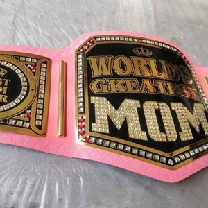 Może przedstawiać: Różowo-złoty pas mistrzowski z kryształkami. Płytka centralna ma czarne tło z złotym napisem "World's Greatest Mom". Płytka boczna ma czarne tło z złotym napisem "Best Mom Ever".
