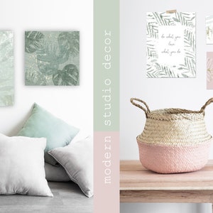 May include: A modern studio decor scene with two green and gold leaf print canvas wall art pieces, three framed prints with a leaf design and inspirational text, a pink and natural woven basket, and a light wood table.
