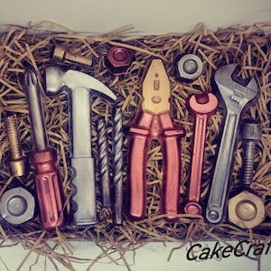 May include: A box filled with brown packing material and a variety of chocolate tools, including a hammer, screwdriver, pliers, wrench, and drill bit. The tools are all brown and silver in colour. The box is labelled "CakeCraft".