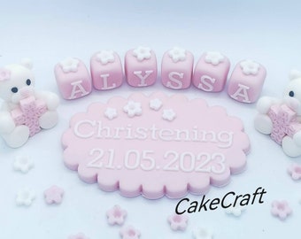 Peut inclure: Décor de gâteau en fondant rose et blanc avec deux ours en peluche tenant des croix, cinq blocs orthographiant "ALYSSA" et un ovale rose avec le texte "Baptême 21.05.2023" et "CakeCraft".