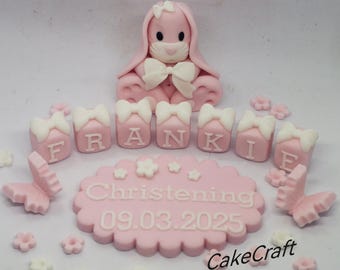 Peut inclure: Un topper de gâteau en forme de lapin rose en fondant avec un nœud blanc, posé sur un gâteau rose avec des lettres en fondant blanc qui épellent "FRANKIE" et une plaque en fondant rose avec le texte "Christening 09.03.2025" et le texte "CakeCraft".