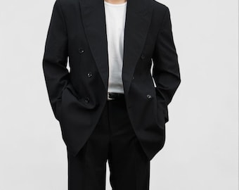 Puede incluir: Un blazer negro de doble botonadura con pantalones negros a juego. El blazer tiene un diseño clásico con solapas de muesca y varios botones. Se lleva una camiseta blanca debajo. El conjunto es de estilo formal.