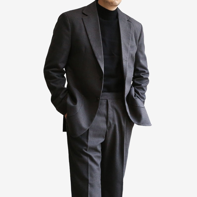 K&ouml;nnte beinhalten: Ein dunkelgrauer Anzug mit einem einreihigen Sakko und einer passenden Hose. Das Sakko hat einen Schalkragen und zwei Pattentaschen. Die Hose hat einen flachen Vorderteil und ein gerades Bein.