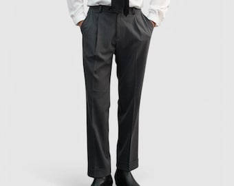 May include: Charcoal gray pleated trousers with a cuffed hem. The pants are paired with black leather boots and a white dress shirt with a black tie. The model is standing with their hands in their pockets.