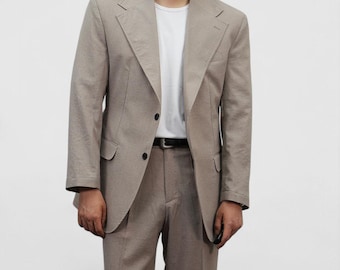 Puede incluir: Un traje marrón claro compuesto por una americana y un pantalón. La americana tiene dos botones negros y solapas con muescas. También se ve una camiseta blanca y un cinturón negro.