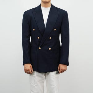 May include: A navy blue double-breasted blazer with gold-colored buttons. The blazer has a classic design with a notched lapel and two front pockets. The person is wearing a white t-shirt and white pants.