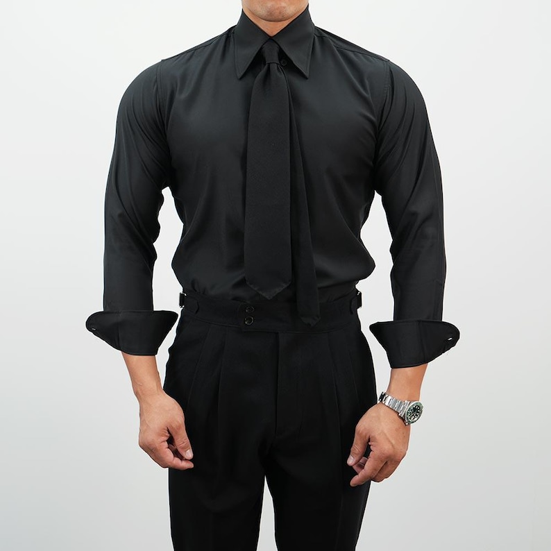 May include: A black dress shirt with a matching tie, paired with black pleated trousers. The shirt features long sleeves with buttoned cuffs. A silver wristwatch is visible on the wrist.
