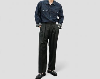May include: A person wearing a dark denim button-down shirt with two chest pockets and black pleated trousers. The person is also wearing black loafers with gold accents.