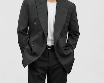 Puede incluir: Un blazer gris oscuro de doble botonadura con una camiseta blanca debajo. El blazer tiene solapas con muescas y un corte clásico. El modelo también lleva pantalones negros y un cinturón negro.