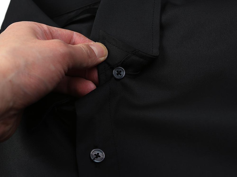 May include: Close-up of a black button-down shirt. The image focuses on the collar and buttons, with a hand partially visible. The shirt is made of a smooth, dark fabric.