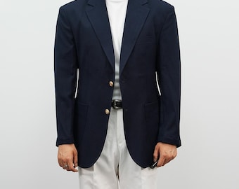 Puede incluir: Una blazer azul marino con botones dorados, usada sobre una camiseta blanca y combinada con pantalones blancos. La blazer tiene un diseño clásico con solapas con muescas y cierre de dos botones. Se ve un cinturón negro.
